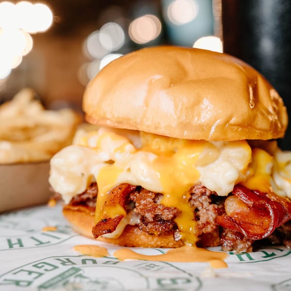 Close-up of a burger with mac and cheese, bacon, and cheese sauce, with fries in the background.