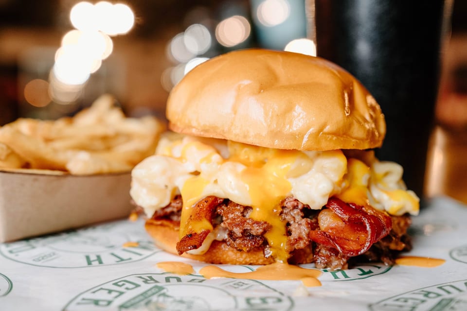Close-up of a burger with mac and cheese, bacon, and cheese sauce, with fries in the background.