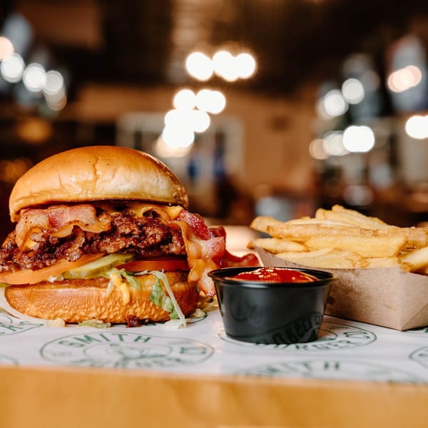 A bacon cheeseburger with fries and ketchup on a branded paper, with blurred lights in the background.
