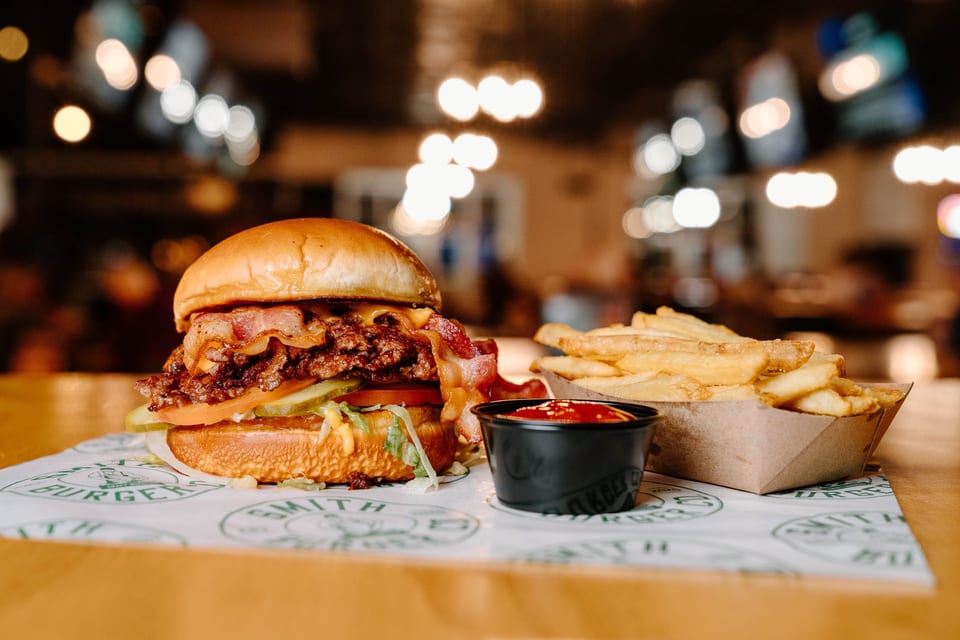 A bacon cheeseburger with fries and ketchup on a branded paper, with blurred lights in the background.
